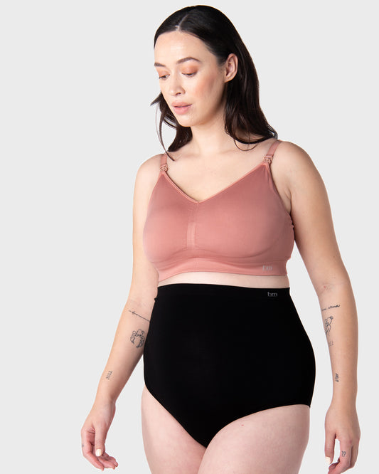Pregnant Woman wearing a pink nursing bra and black high-waisted underwear on a white background