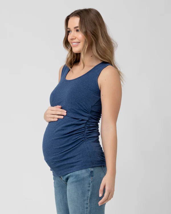 Pregnant Woman wearing a blue nursing tank top and jeans, standing against a plain background