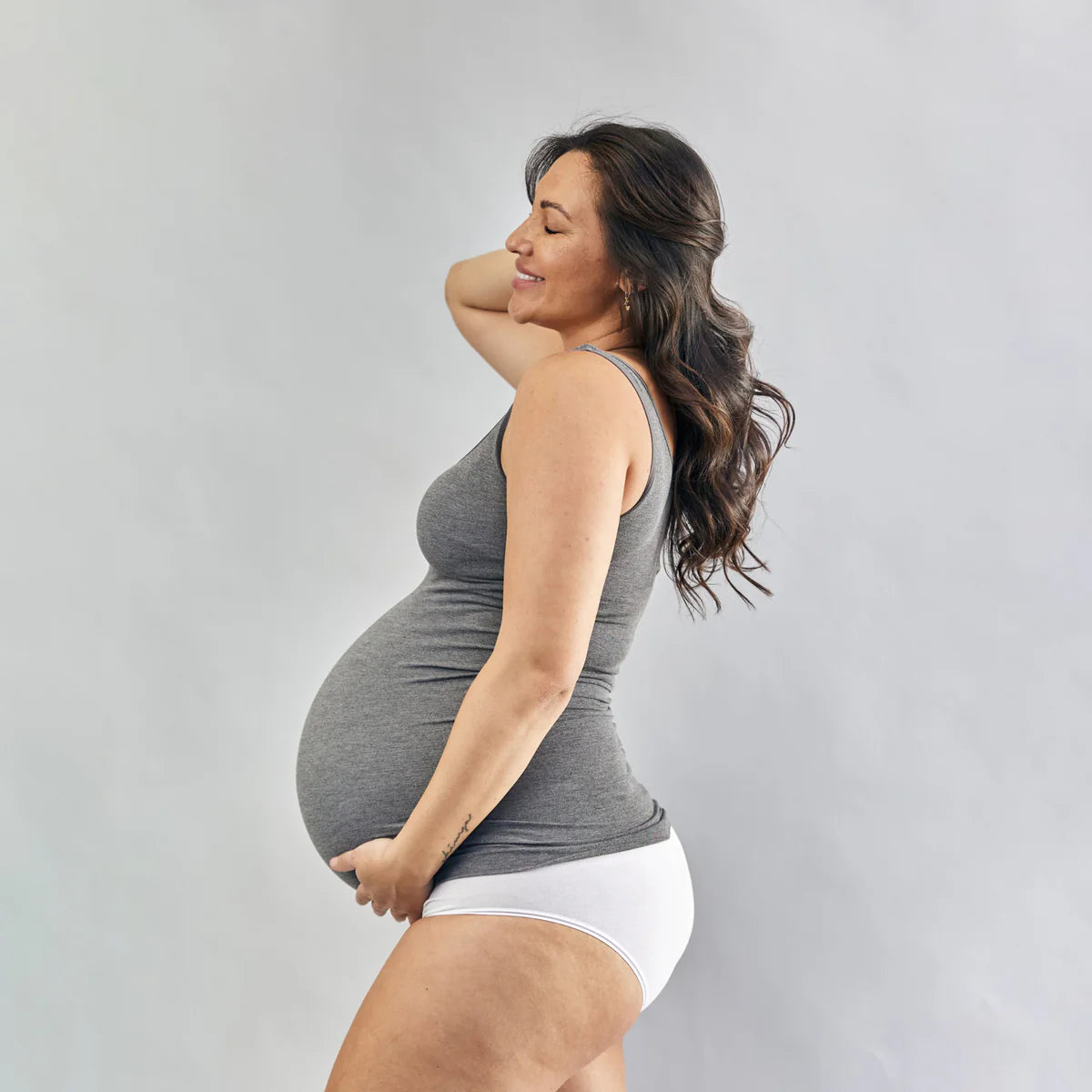 Pregnant woman wearing a grey tank top and white underwear against a grey background