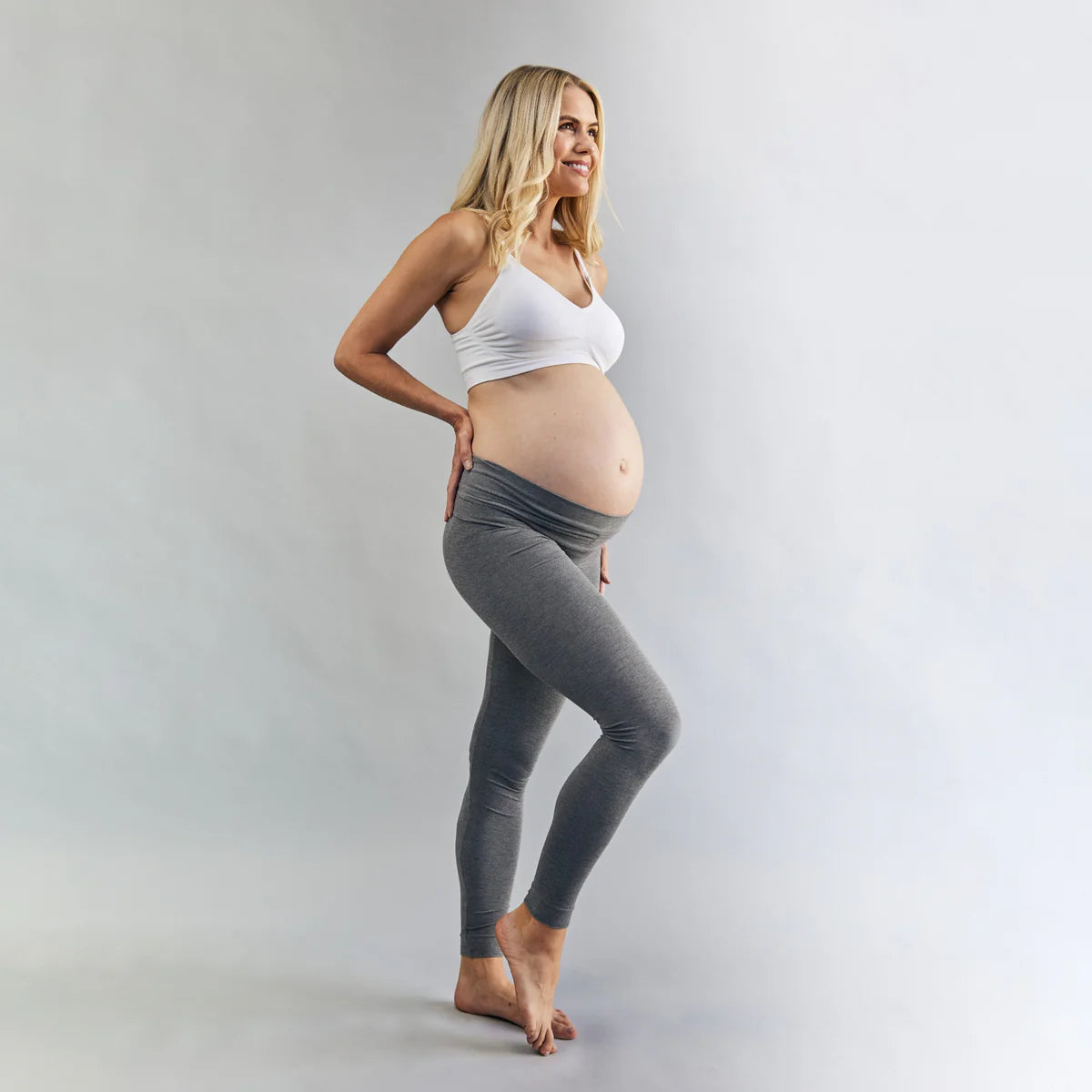 Pregnant woman wearing grey leggings and a white top on a plain background