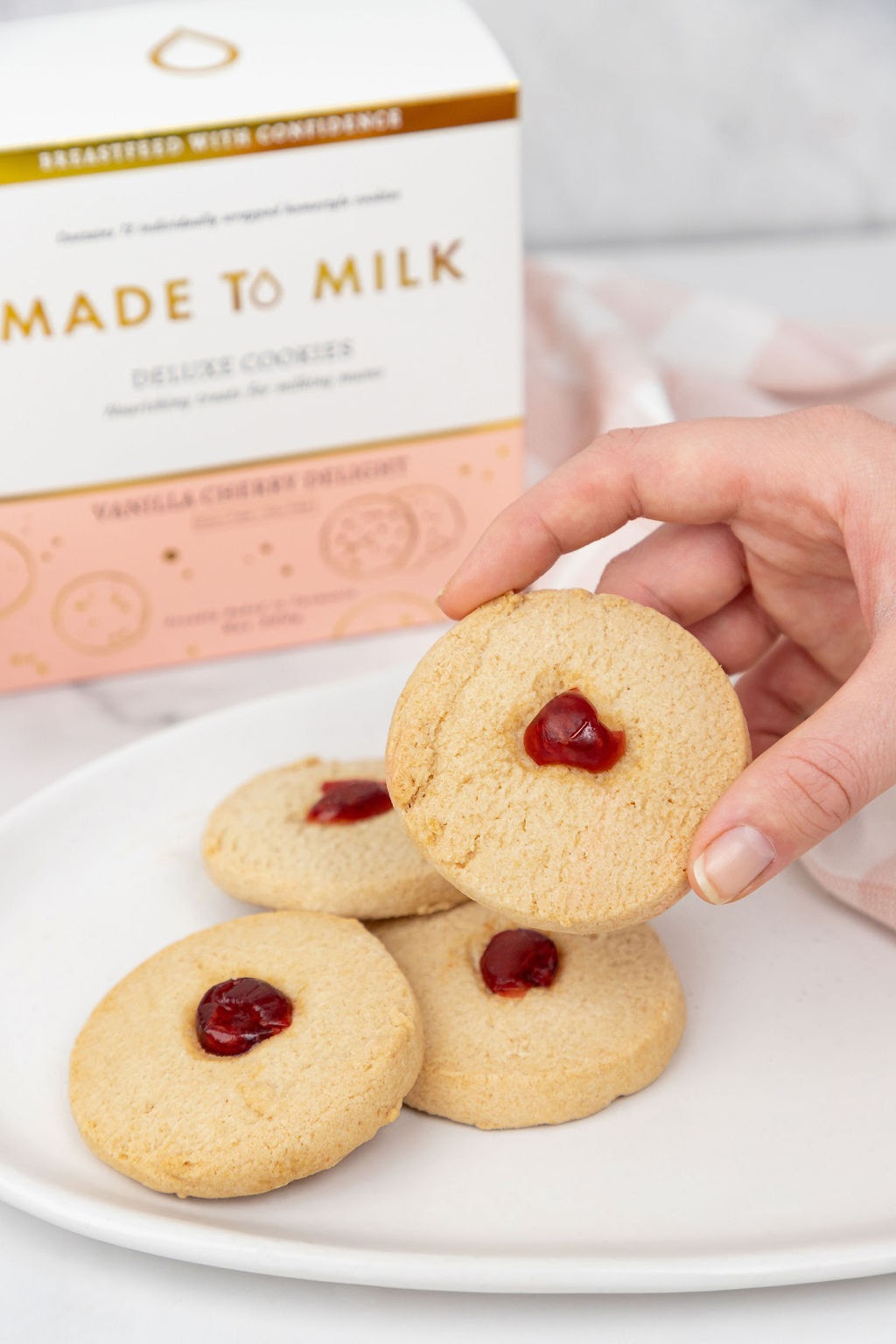 Person holding a cookie with jam filling on a plate with 'Made to Milk' cookies in the background.