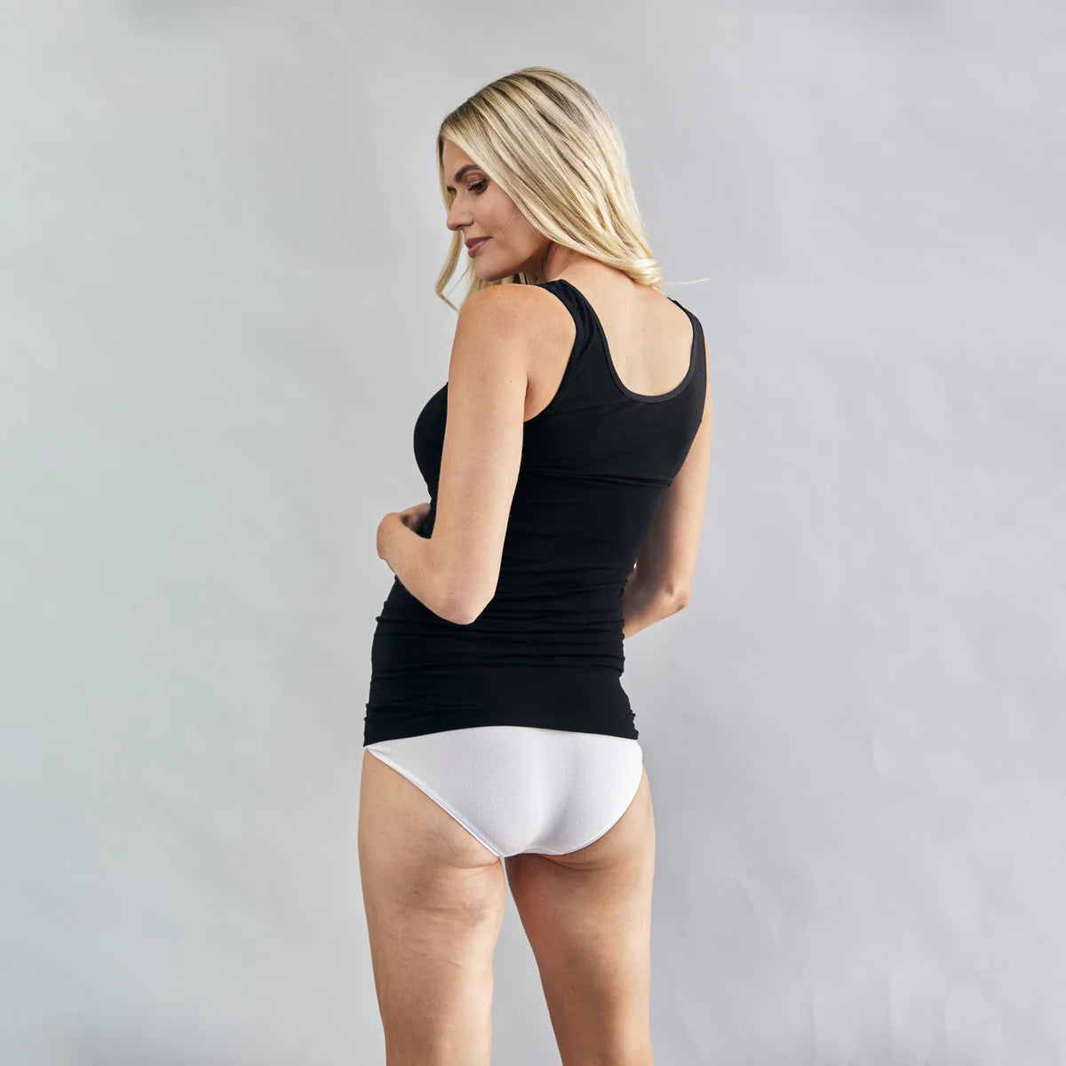 Pregnant Woman wearing a black nursing tank top and white underwear on a plain background