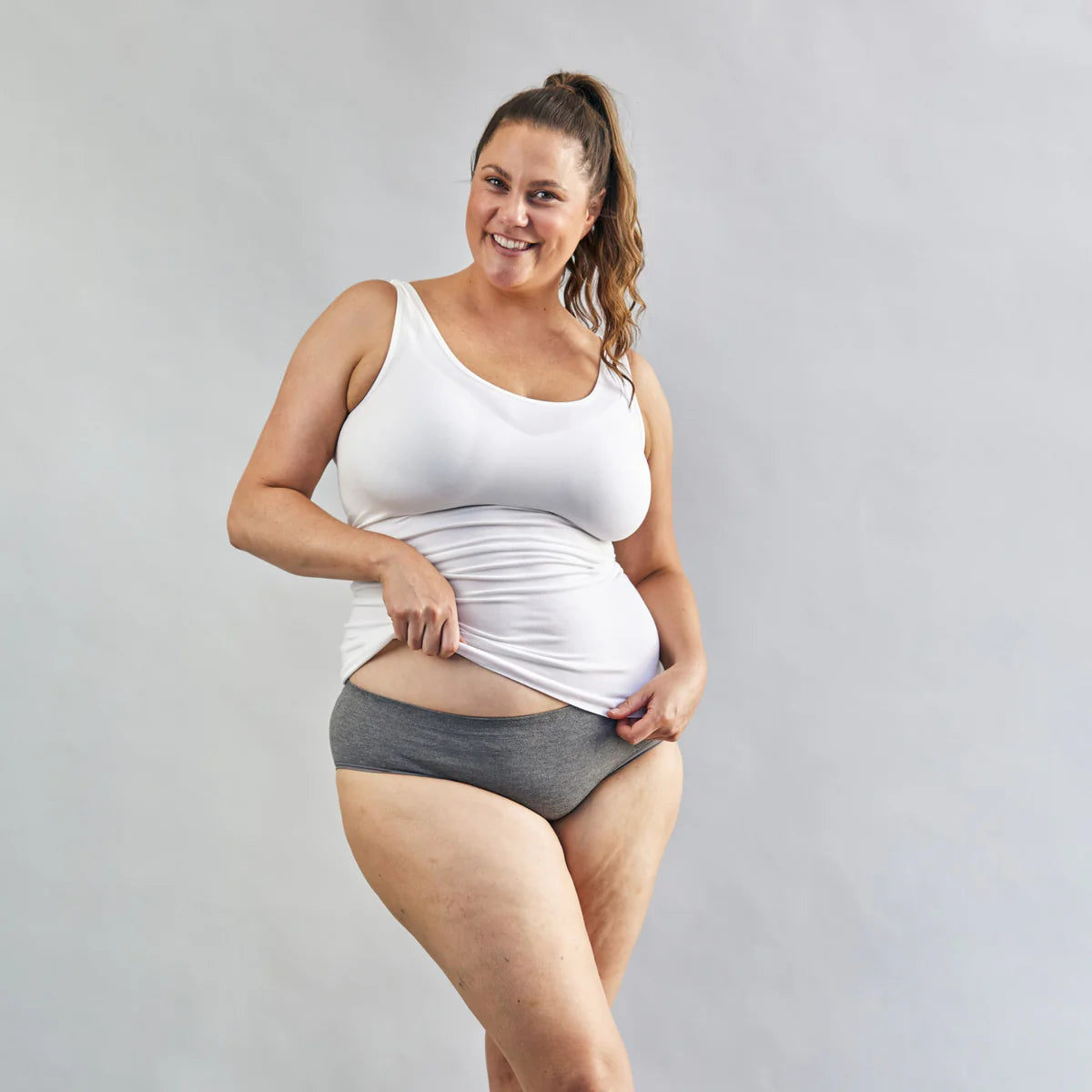 Pregnant Woman wearing a white nursing tank top and grey underwear on a plain background
