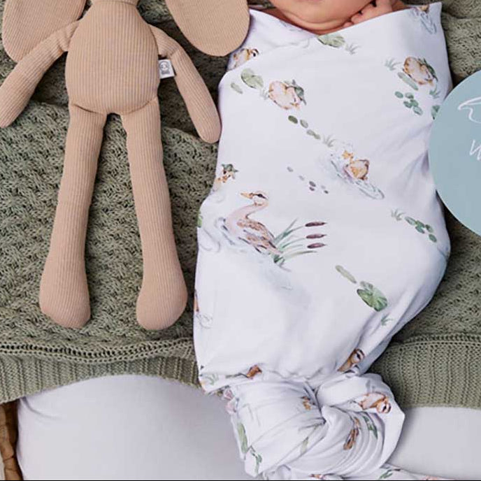 Newborn baby wrapped in a floral swaddle with a hat, lying on a green blanket next to a plush toy and 'hello world' sign.