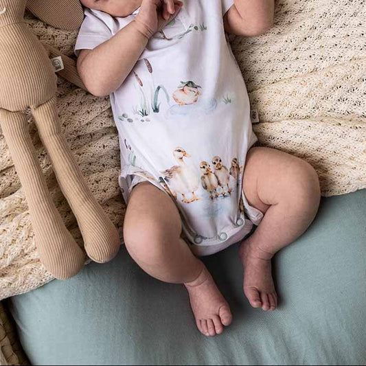 Baby lying on a cushion with a plush toy, wearing a white romper with animal prints and a headband.