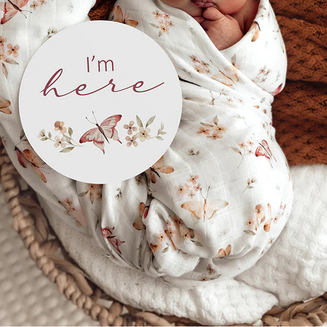 Newborn baby wrapped in a floral blanket with a 'I'm here' sign, lying in a woven basket.