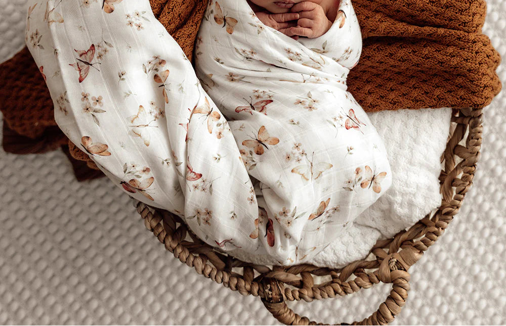 Newborn baby wrapped in a floral blanket with a brown wrap, lying in a wicker basket on a textured surface.