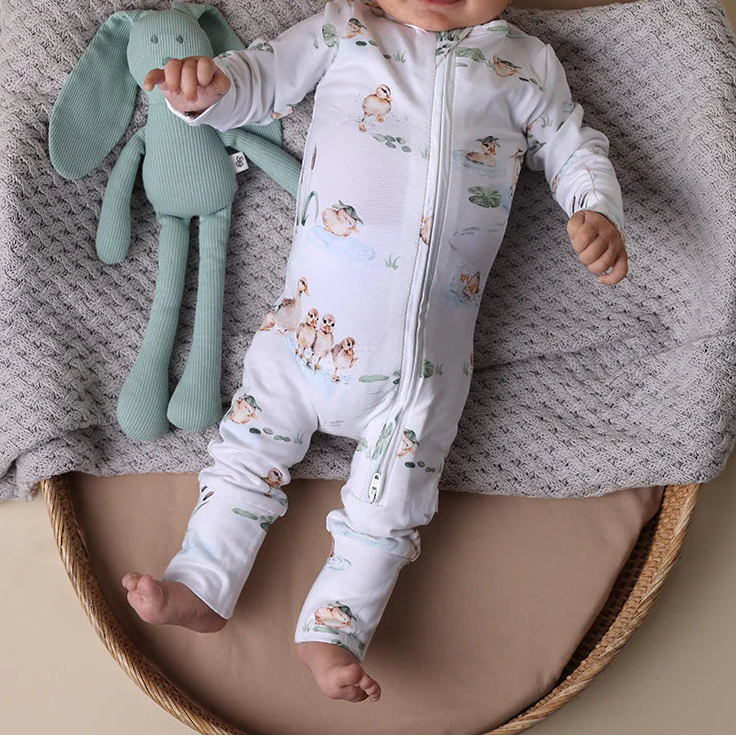 Baby lying on a textured blanket with a green plush toy, wearing a patterned onesie and white cap.