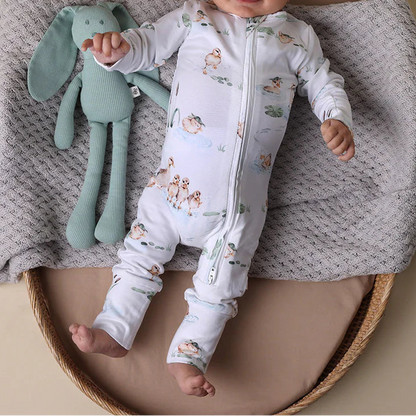 Baby lying on a textured blanket with a green plush toy, wearing a patterned onesie and white cap.