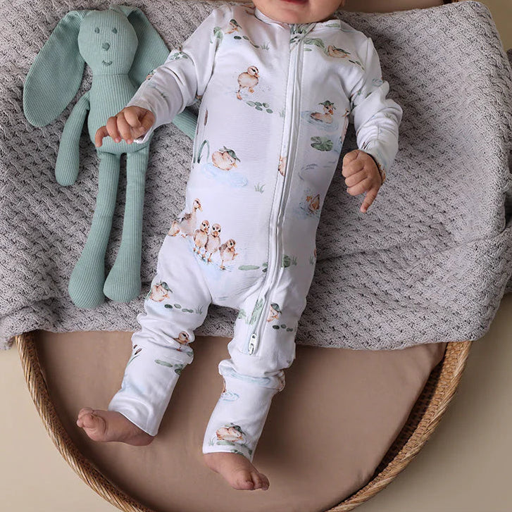 Baby in a white onesie with animal patterns lying on a textured blanket next to a green plush toy.