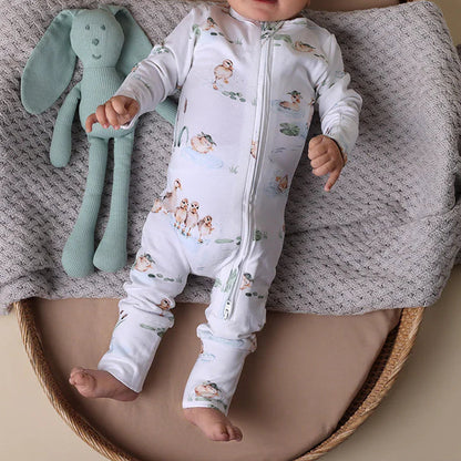 Baby in a white onesie with animal patterns lying on a textured blanket next to a green plush toy.