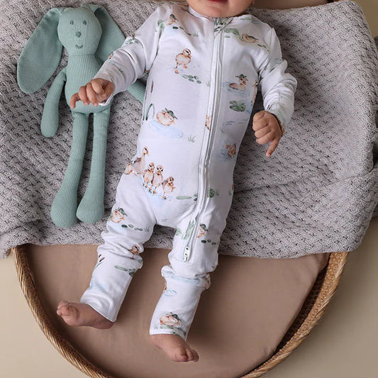 Baby in a white onesie with animal patterns lying on a textured blanket next to a green plush toy.