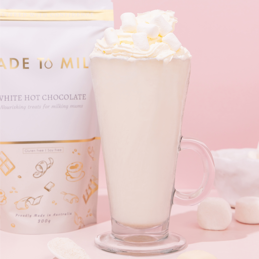 Glass of white hot chocolate with whipped cream next to a product packaging on a pink background